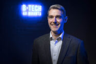 A photo of Mark Walker in a grey suit and a pale blue button-up shirt, standing in front of a neon sign which says B-Tech AV Mounts