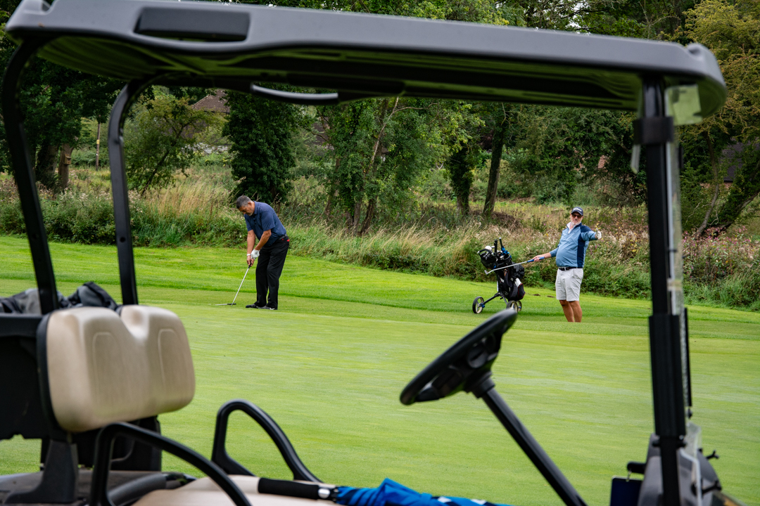 An image of one man facing toward the camera on a Golfing green holding a putter in the air, Whilst another man on the hits a golf ball with a putter
